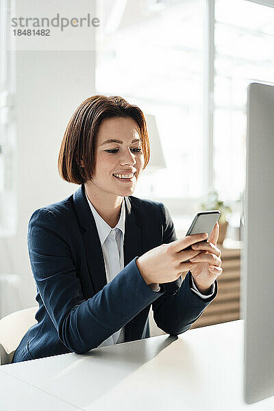 Glückliche Geschäftsfrau sitzt am Schreibtisch im Büro und nutzt ihr Smartphone