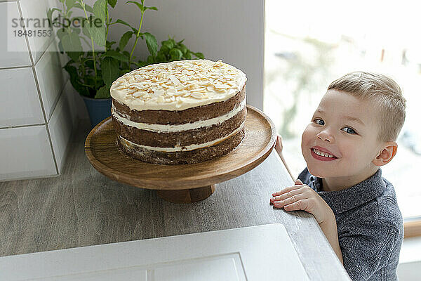 Happy cute boy standing by cake in kitchen