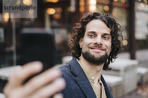 Lächelnder junger Geschäftsmann  der vor dem Café ein Selfie macht