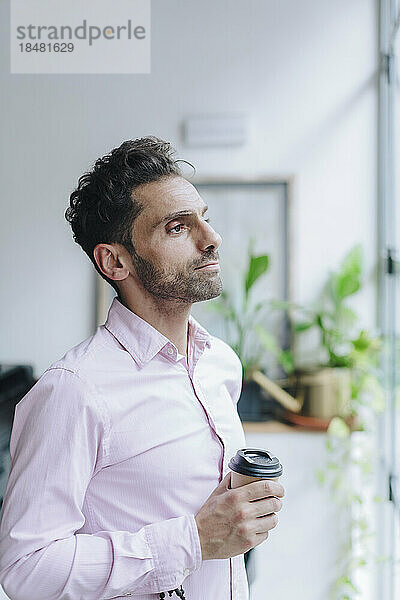 Nachdenklicher reifer Geschäftsmann steht mit Einwegkaffeetasse im Büro