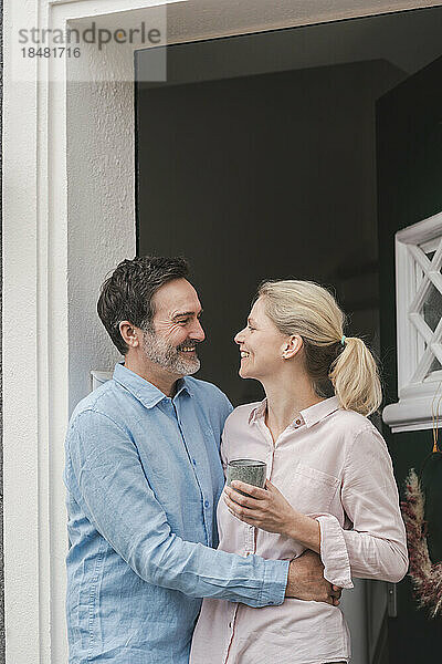 Glücklicher Mann umarmt Frau mit Tasse am Eingang des Hauses