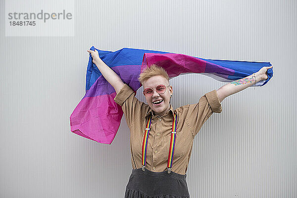 Glückliche nicht-binäre Person mit bisexueller Flagge vor weißer Wand