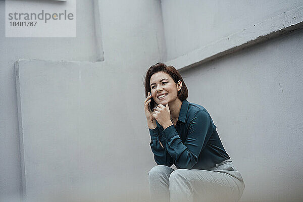 Lächelnde Geschäftsfrau  die vor einer grauen Wand über ihr Mobiltelefon spricht
