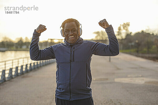 Glücklicher älterer Mann mit Kopfhörern  der an der Promenade seine Muskeln spielen lässt