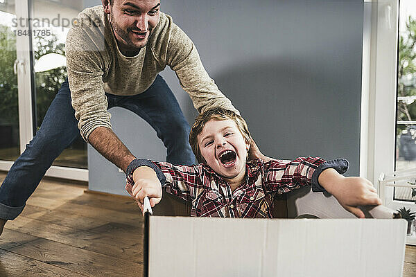 Fröhlicher Junge mit Vater  der einen Karton schiebt und zu Hause genießt