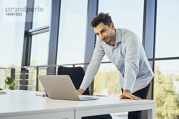 Lächelnder Geschäftsmann beobachtet Laptop am Schreibtisch im Büro