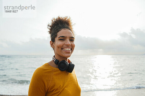 Glückliche Frau mit kabellosen Kopfhörern steht am Strand