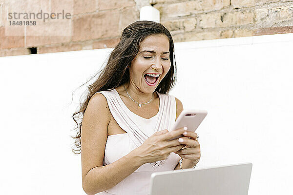 Fröhliche Frau sitzt mit Smartphone vor der Wand