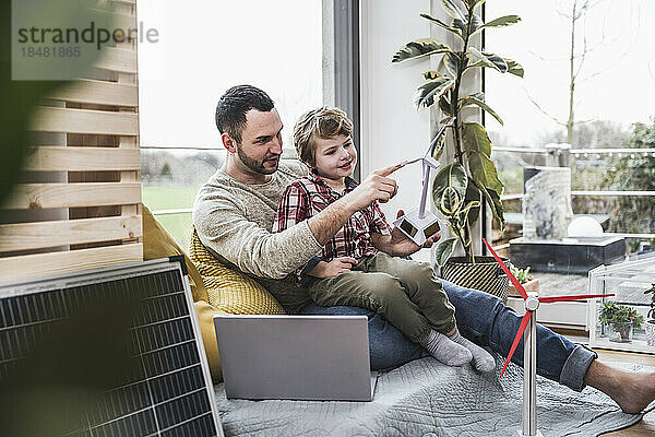 Vater berührt Modell einer Windkraftanlage  Sohn sitzt zu Hause