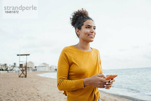 Nachdenkliche junge Frau mit Smartphone steht am Strand