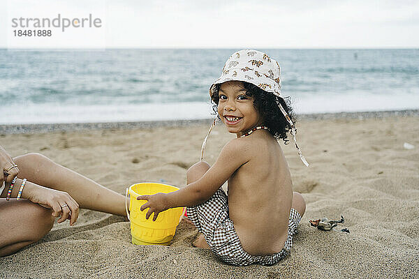 Fröhlicher süßer Junge mit Hut  der mit Eimer im Sand spielt