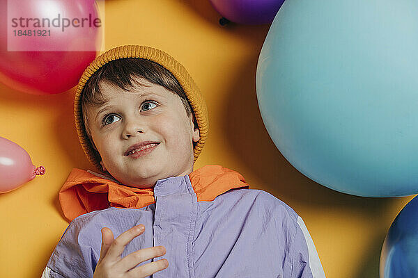 Smiling boy lying amidst multi colored balloons against yellow background