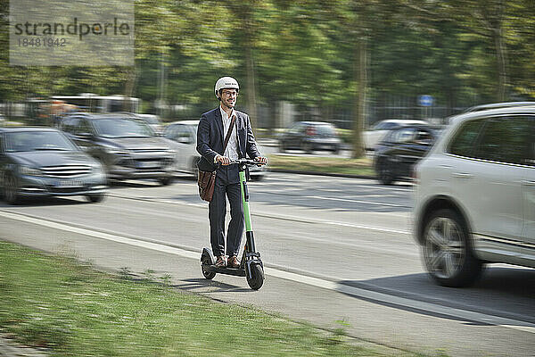 Young businessman wearing helmet riding electric push scooter on road