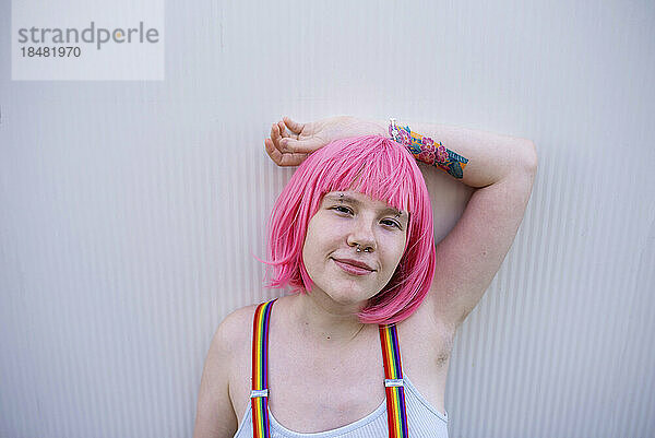 Lächelnde nicht-binäre Person mit rosa Perücke vor grauer Wand