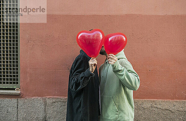 Schwules Paar bedeckt Gesicht mit rotem herzförmigem Ballon