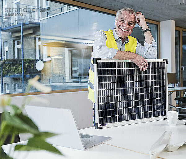 Lächelnder leitender Ingenieur mit Solarpanel auf dem Schreibtisch im Büro