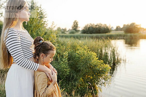 Nachdenkliches Mädchen steht mit Schwester bei Sonnenuntergang am See