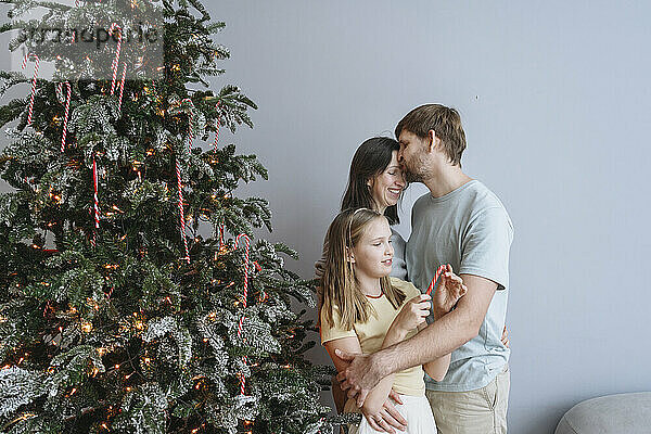 Mann küsst Frau und Tochter zu Hause am Weihnachtsbaum auf die Stirn