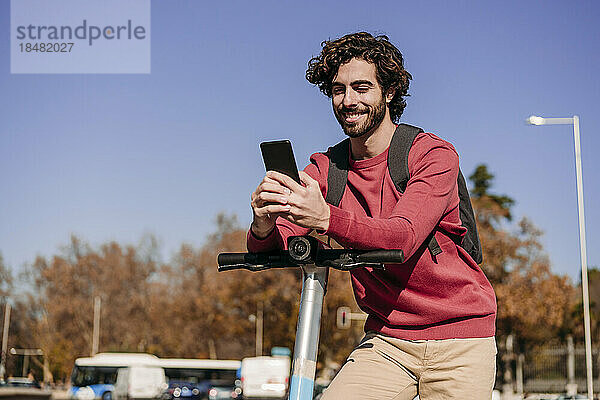 Lächelnder junger Mann stützt sich auf einen Elektroroller und benutzt ein Smartphone