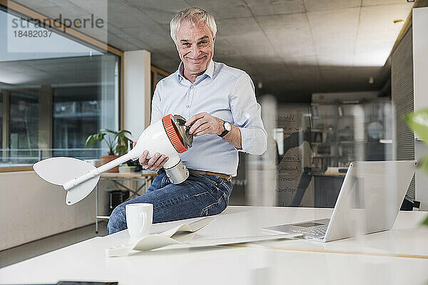 Lächelnder leitender Ingenieur mit Windturbinenrotor sitzt auf dem Schreibtisch im Büro
