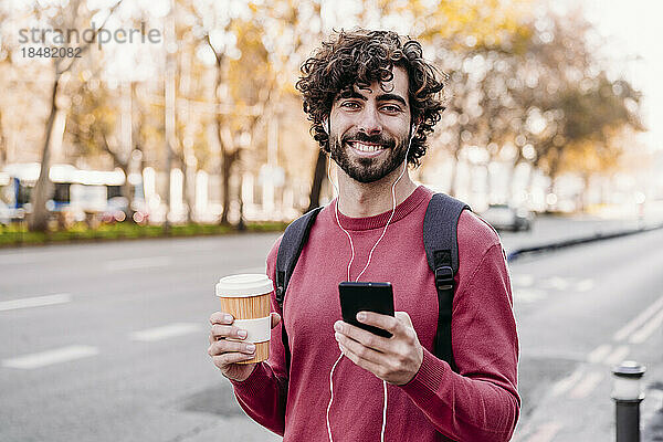 Glücklicher junger Mann mit Handy und Kaffeetasse steht am Fußweg