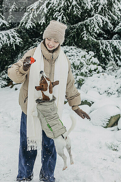 Lächelnde junge Frau spielt mit Hund im verschneiten Park