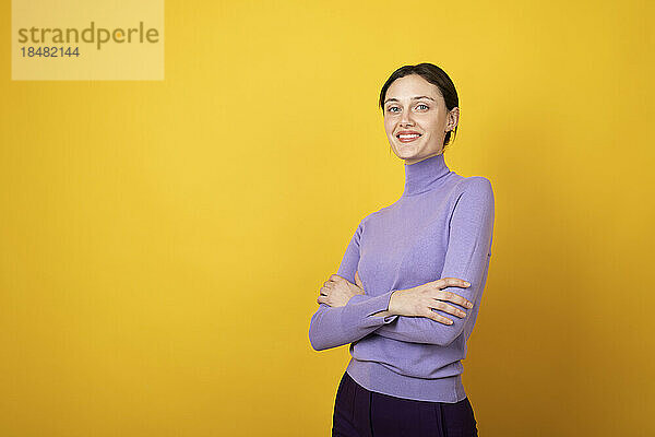 Happy woman standing with arms crossed against yellow background