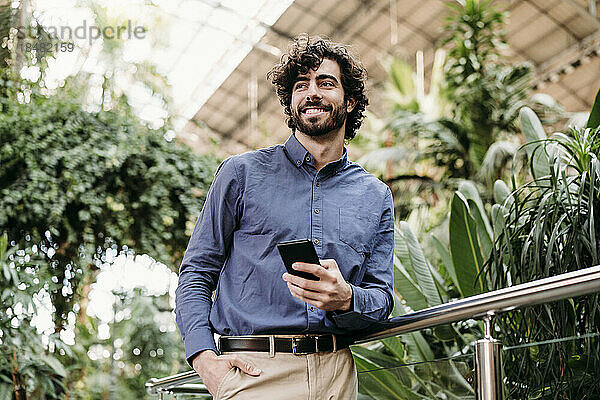 Lächelnder Geschäftsmann mit Smartphone steht am Geländer vor Pflanzen