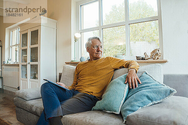 Älterer Mann mit Tablet-PC sitzt zu Hause auf dem Sofa im Wohnzimmer
