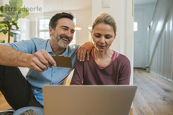 Glücklicher Ehemann hält Kreditkarte in der Hand  während Frau zu Hause Laptop benutzt