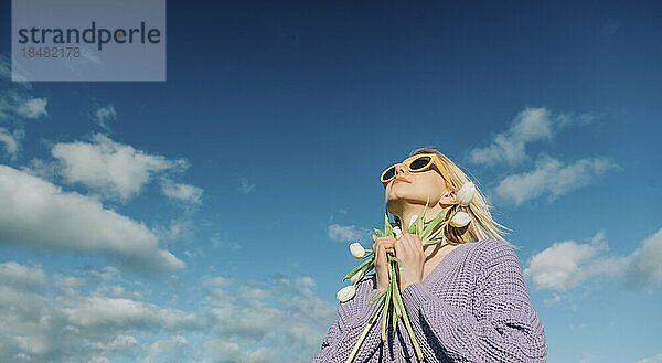 Frau mit Sonnenbrille steht mit Tulpen unter blauem Himmel