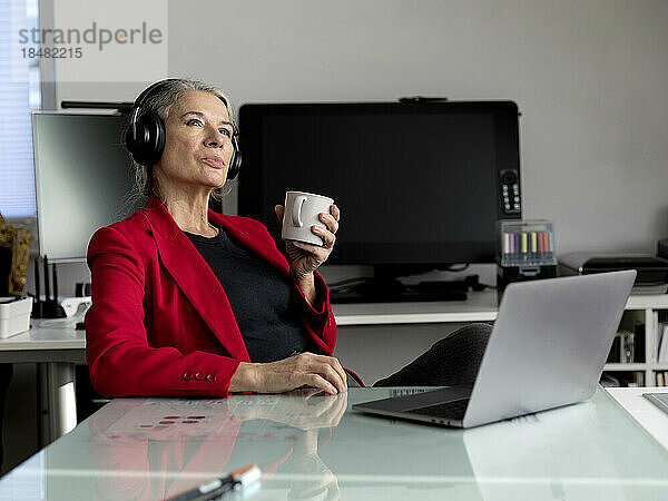Lächelnde ältere Geschäftsfrau mit Kaffeetasse sitzt im Büro