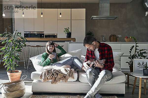 Mann spielt Ukulele  sitzt neben Frau und benutzt Smartphone auf der heimischen Couch