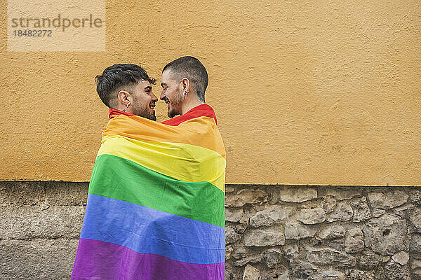 Glückliches schwules Paar  bedeckt mit Regenbogenfahne an der Wand