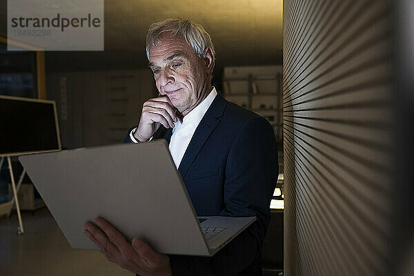 Nachdenklicher Geschäftsmann mit der Hand am Kinn  der im Büro auf den Laptop schaut