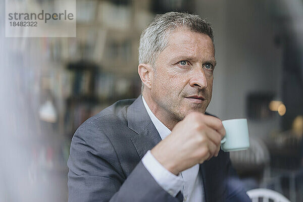 Nachdenklicher Geschäftsmann mit Kaffeetasse durch Glasfenster gesehen