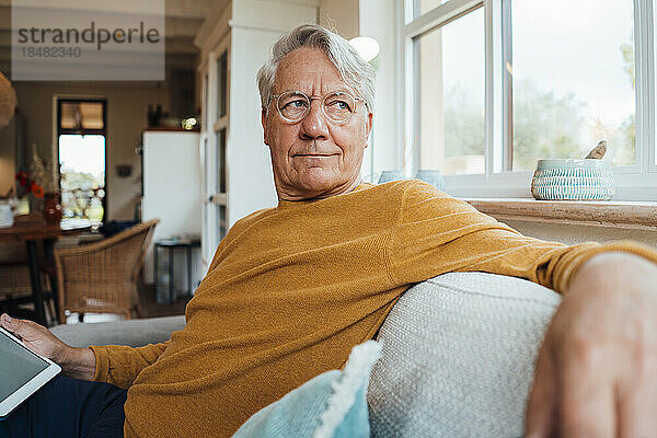 Nachdenklicher älterer Mann sitzt mit Tablet-PC auf dem Sofa im heimischen Wohnzimmer