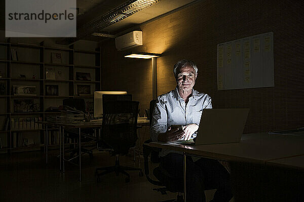 Lächelnder älterer Geschäftsmann sitzt mit Laptop am Schreibtisch im Büro