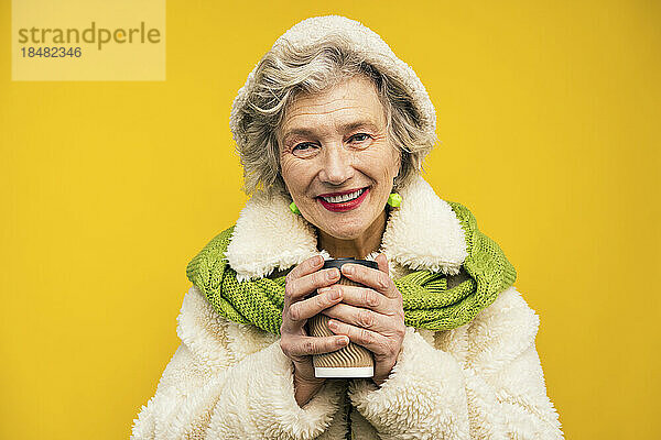 Ältere Frau hält Einwegkaffeetasse vor gelbem Hintergrund