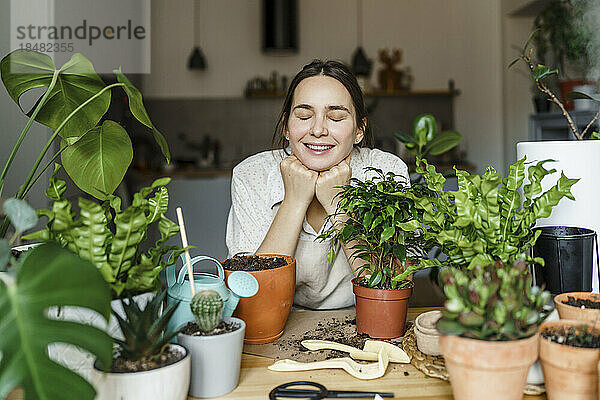 Lächelnde Frau mit geschlossenen Augen  die sich zu Hause neben Pflanzen auf den Tisch lehnt