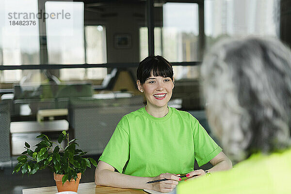 Smiling woman in green t-shirt looking at colleague in office