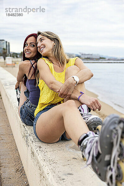 Lächelnde Freunde mit Inline-Skates sitzen Rücken an Rücken an der Promenade