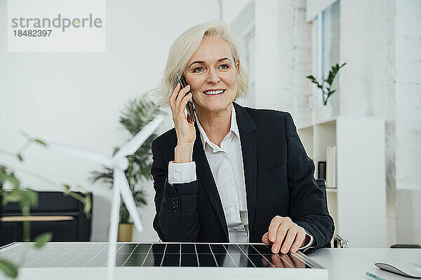 Reife Geschäftsfrau sitzt mit Solarpanel und telefoniert am Schreibtisch
