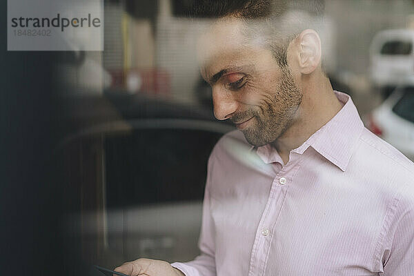 Lächelnder reifer Geschäftsmann mit Smartphone durch Glas gesehen