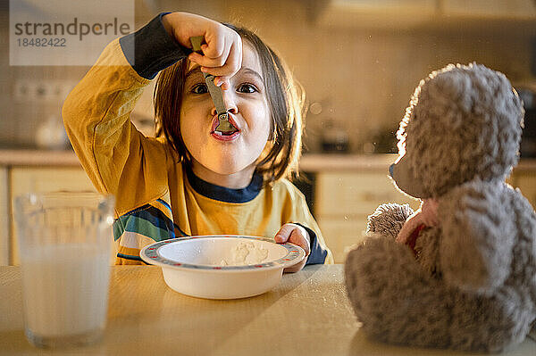 Junge isst Haferbrei zum Frühstück mit Teddybär auf dem heimischen Tisch