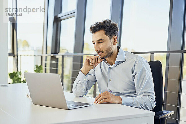 Geschäftsmann mit der Hand am Kinn und Blick auf den Laptop im Büro