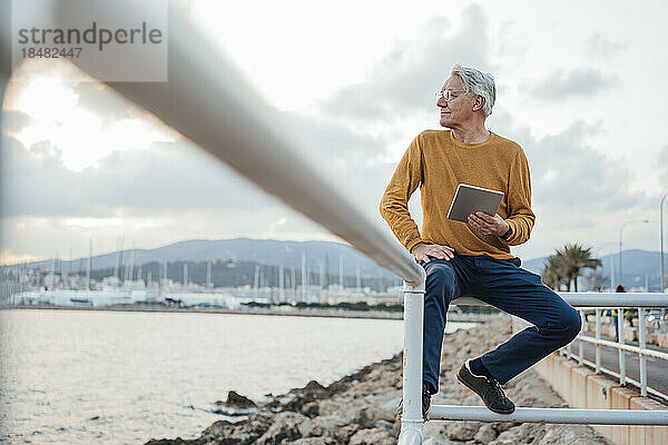Lächelnder älterer Mann sitzt mit Tablet-PC auf dem Geländer