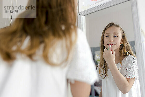 Smiling girl applying lipstick looking in mirror at home