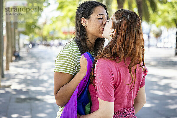 Lesbisches Paar mit Regenbogenfahne küsst sich am Fußweg auf den Mund