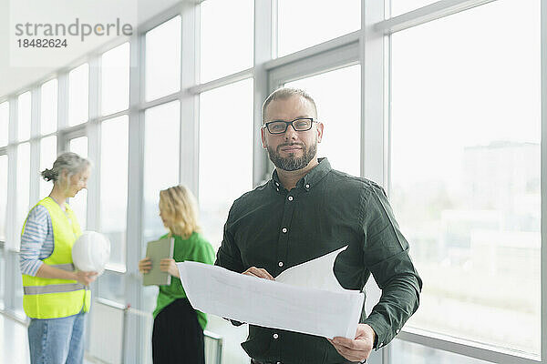 Businessman holding plan on office floor with colleagues in background